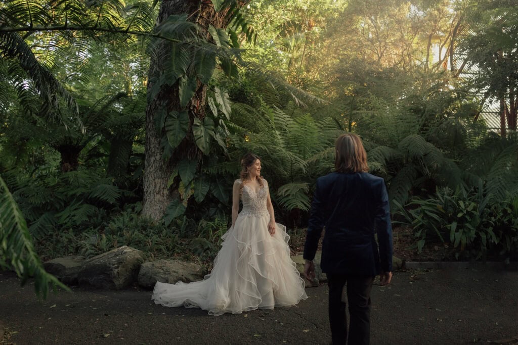 A bride and groom in wedding attire standing in a lush, green forest setting.