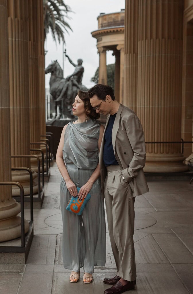 Stylish couple in formal attire standing under classical columns with a statue in the background.